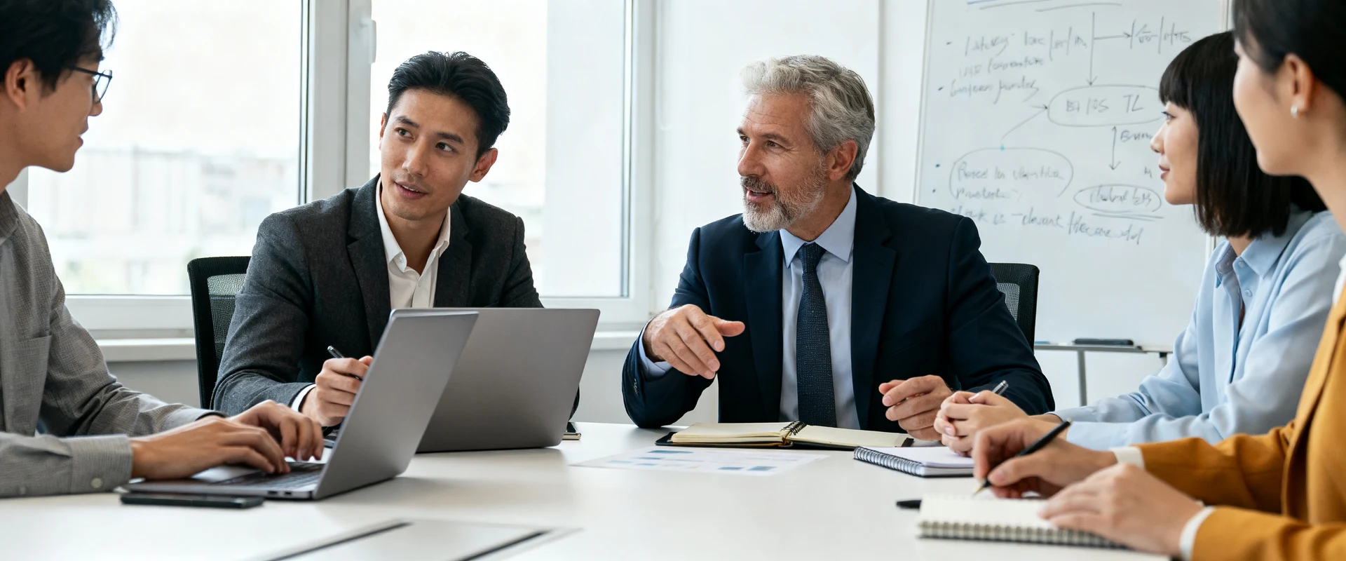 Proposta de valor: o que é, como criar e exemplos práticos Equipe de negócios diversificada colaborando em mesa de conferência moderna com laptops e notebooks, discutindo estratégia em ambiente corporativo autêntico com luz natural e quadro branco ao fundo.